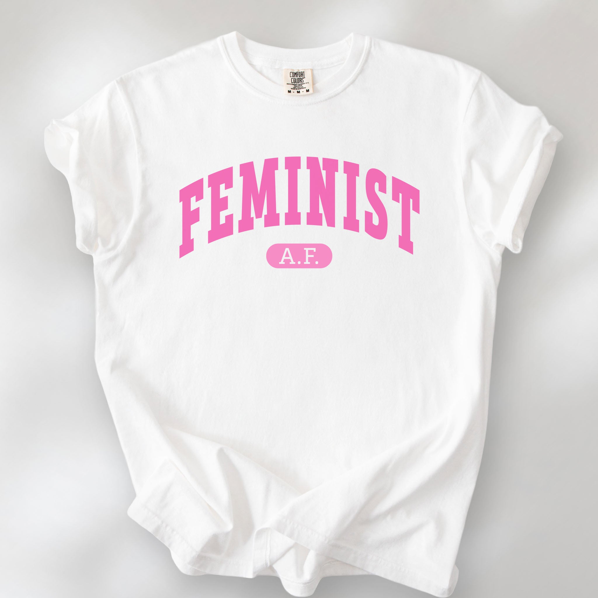 White Comfort Colors tee with pink text that reads “Feminist A.F.” in a collegiate style.