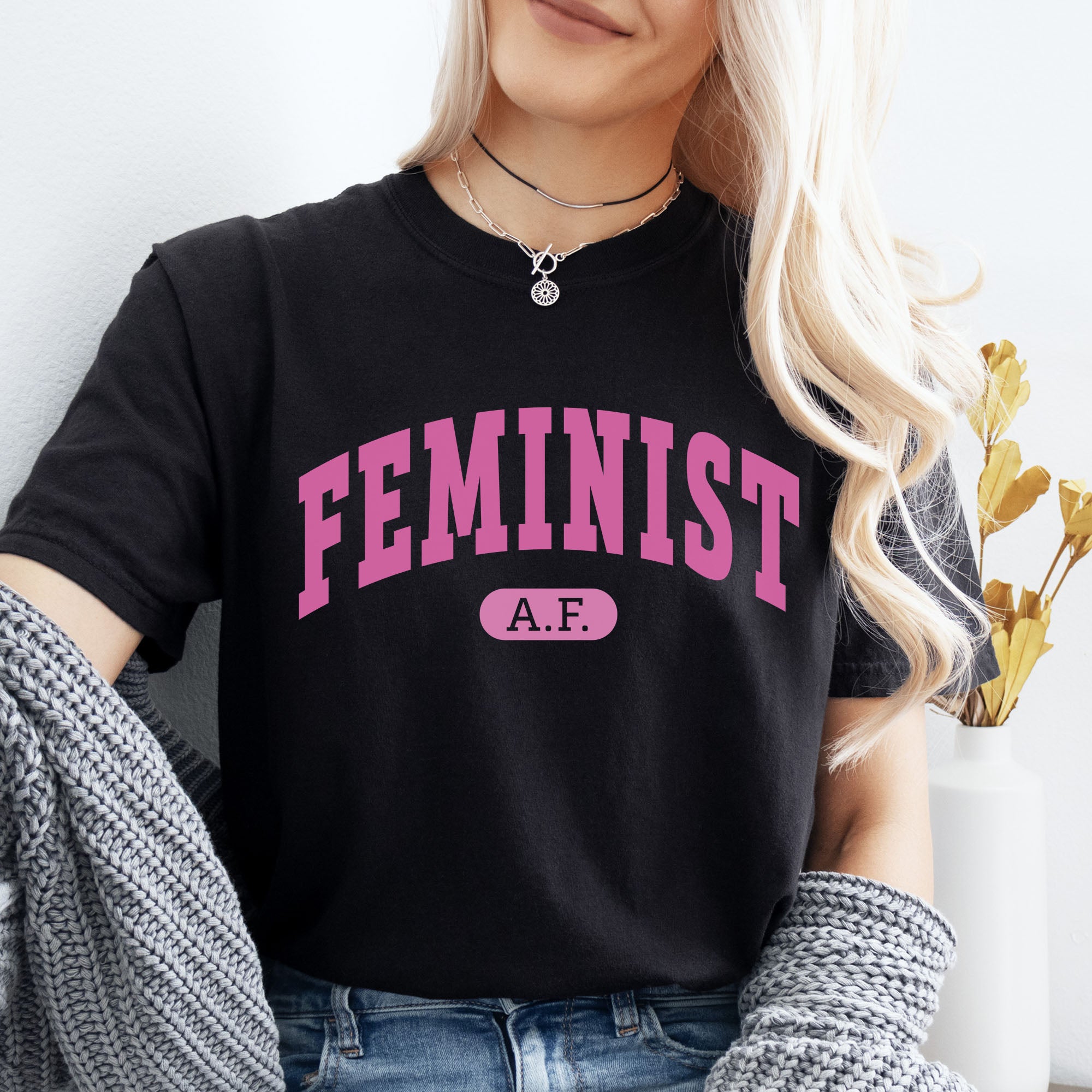 Female model wearing black Comfort Colors tee with pink text that reads “Feminist A.F.” in a collegiate style.