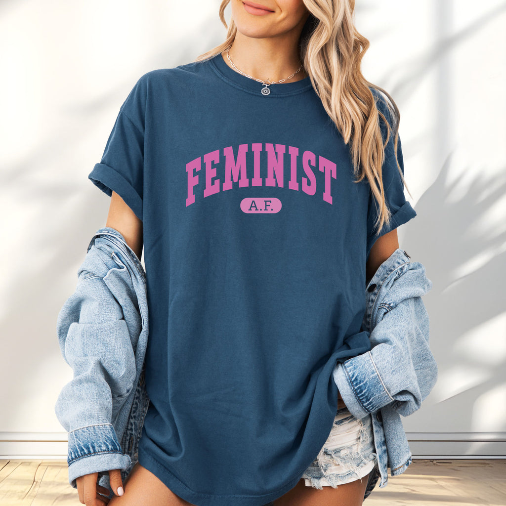 Female model wearing oversized midnight Comfort Colors tee with pink text that reads “Feminist A.F.” in a collegiate style.