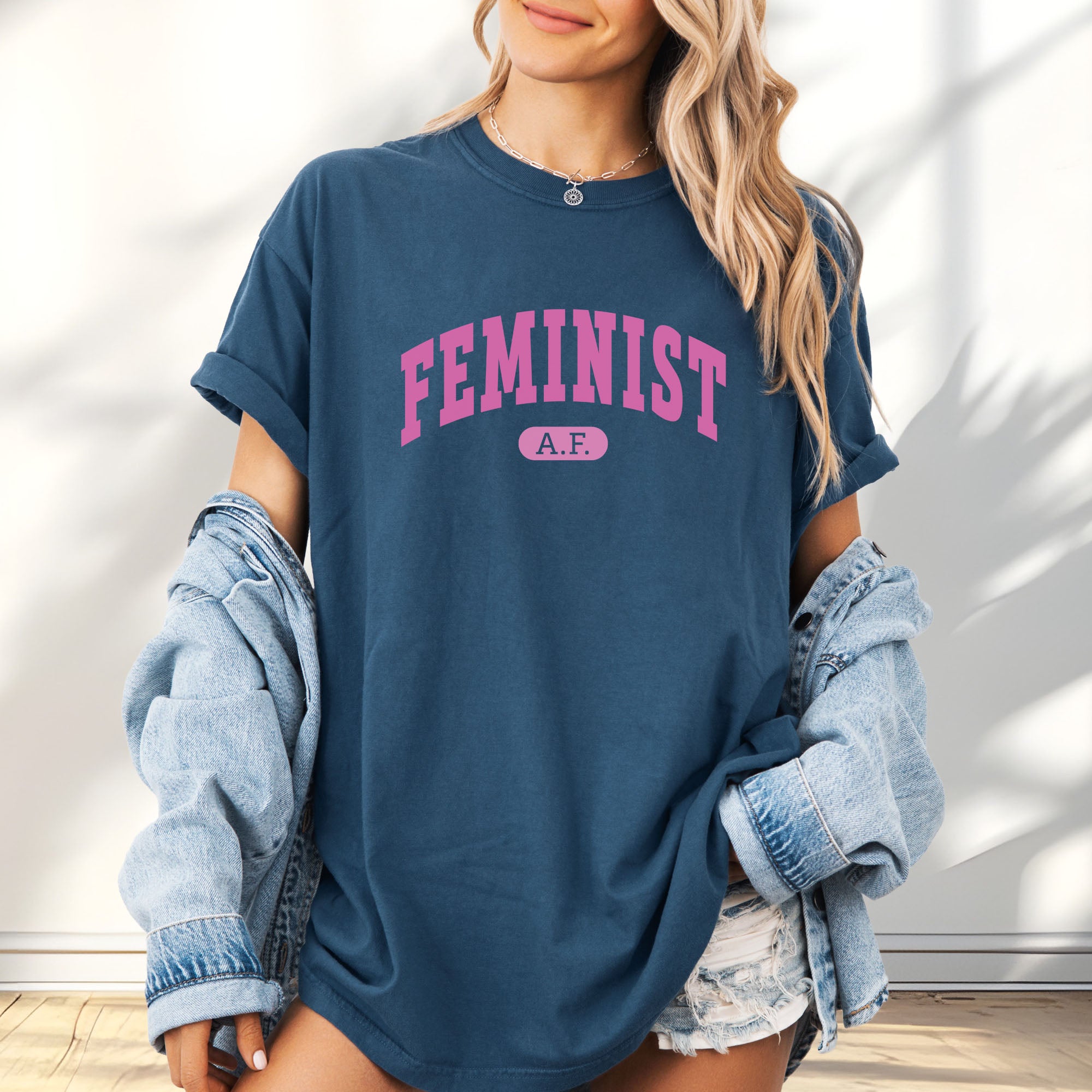 Female model wearing oversized midnight Comfort Colors tee with pink text that reads “Feminist A.F.” in a collegiate style.