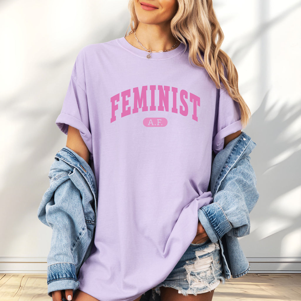 Female model wearing oversized orchid Comfort Colors tee with pink text that reads “Feminist A.F.” in a collegiate style.