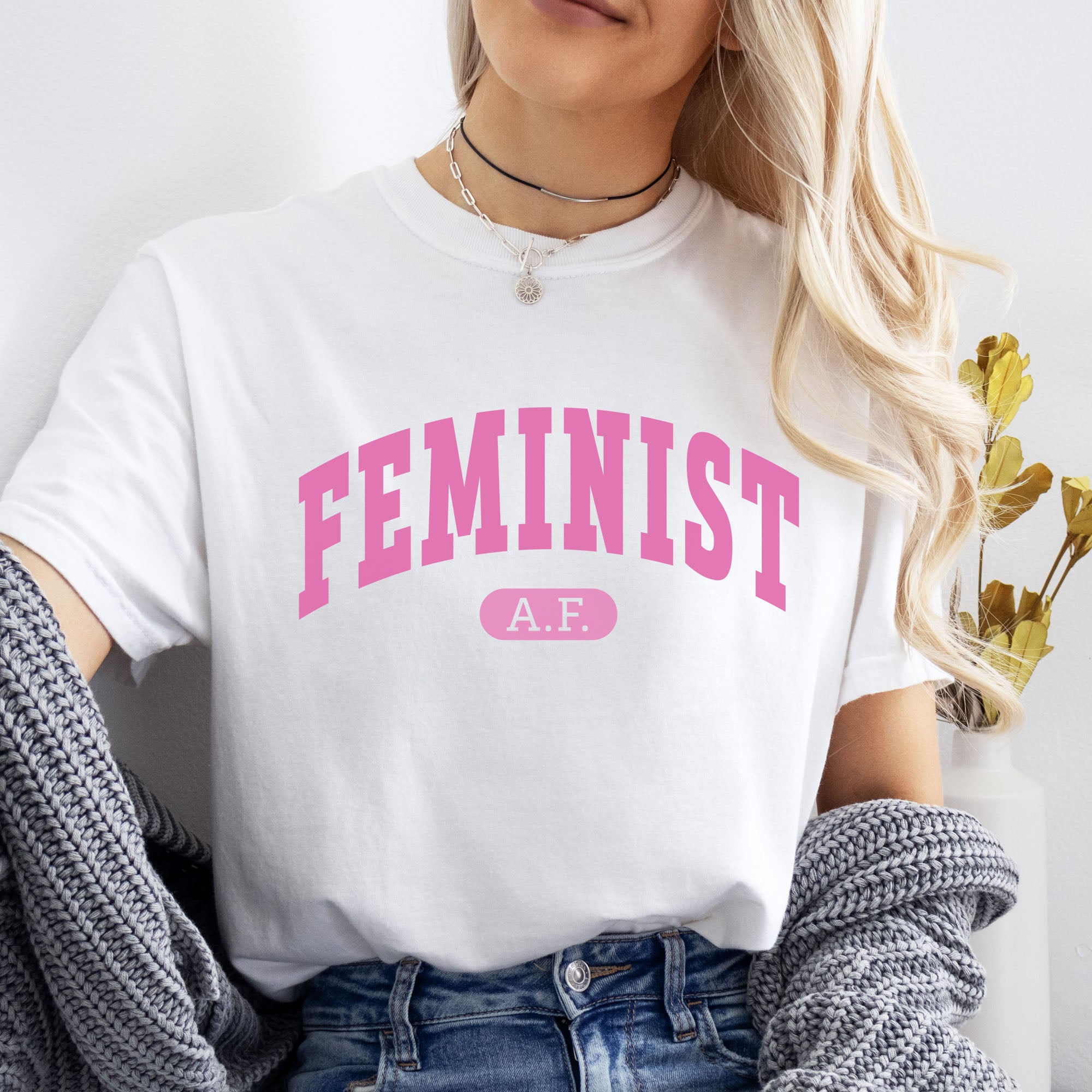 Female model wearing white Comfort Colors tee with pink text that reads “Feminist A.F.” in a collegiate style.