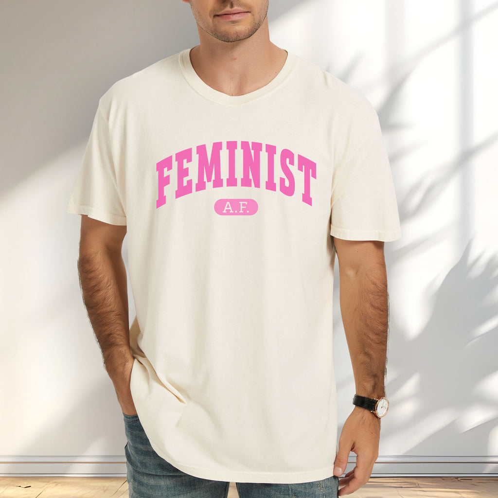 Male model wearing ivory Comfort Colors tee with pink text that reads “Feminist A.F.” in a collegiate style.