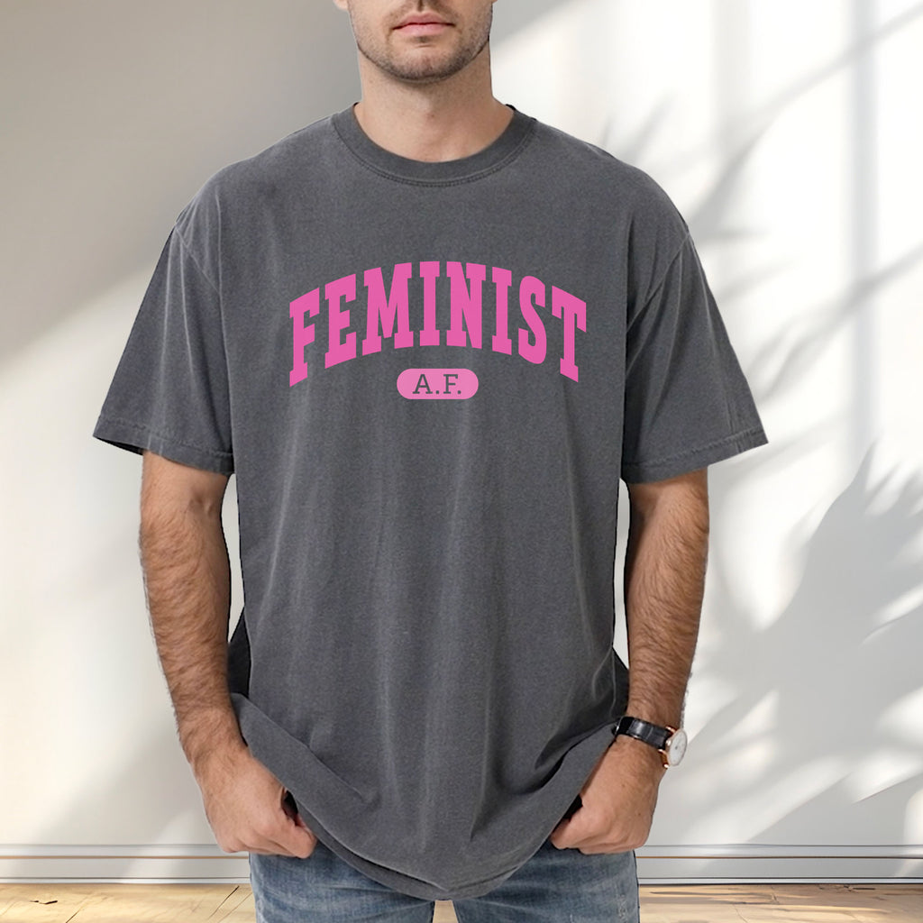 Male model wearing pepper Comfort Colors tee with pink text that reads “Feminist A.F.” in a collegiate style.