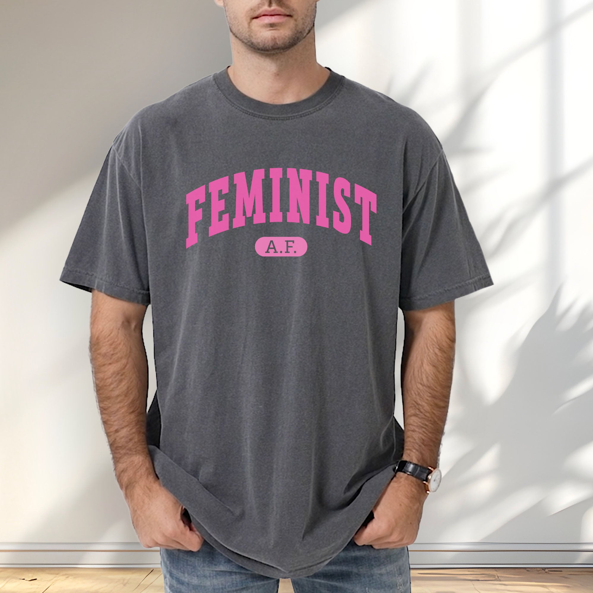 Male model wearing pepper Comfort Colors tee with pink text that reads “Feminist A.F.” in a collegiate style.