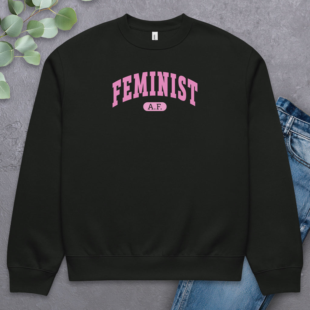 Flat black Bella + Canvas heavyweight sweatshirt with pink text that reads “Feminist A.F.” in a collegiate style, styled with jeans and greenery.