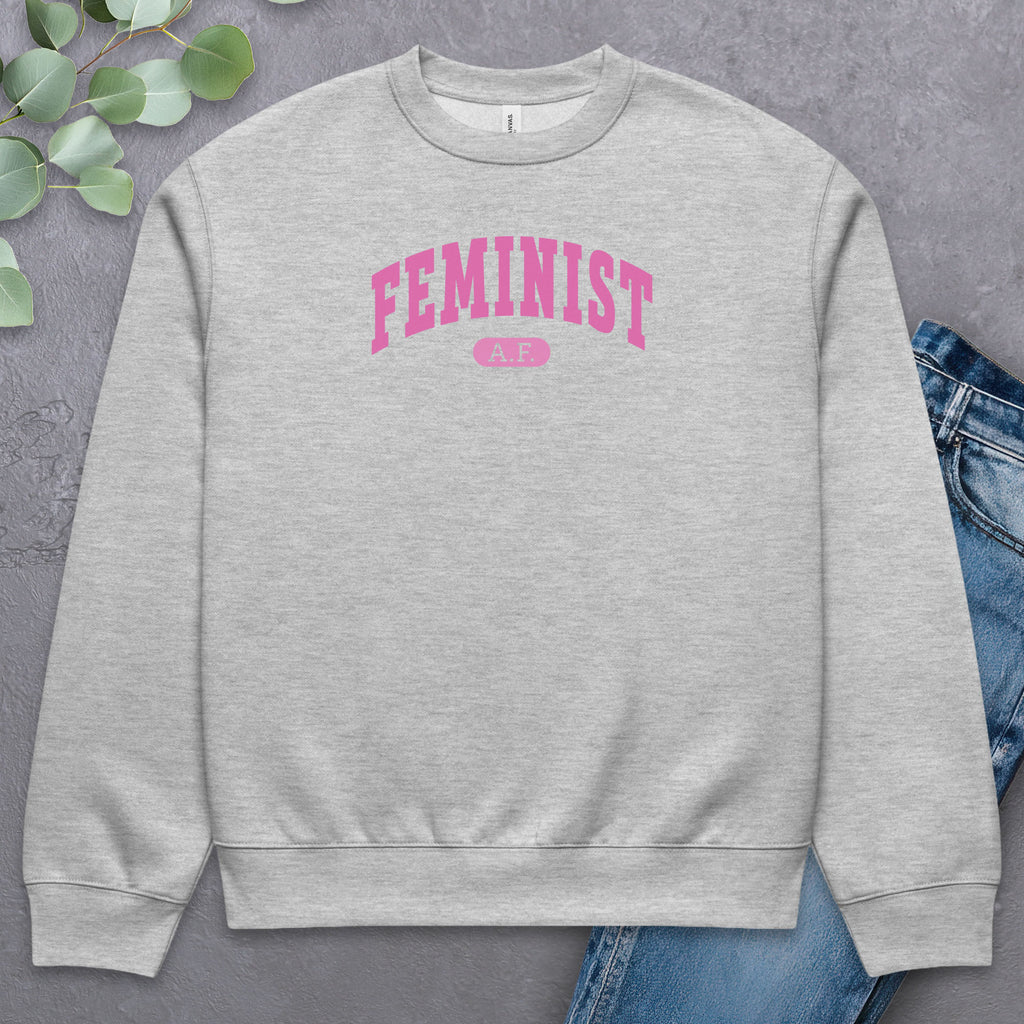 Flat grey Bella + Canvas heavyweight sweatshirt with pink text that reads “Feminist A.F.” in a collegiate style, styled with jeans and greenery.