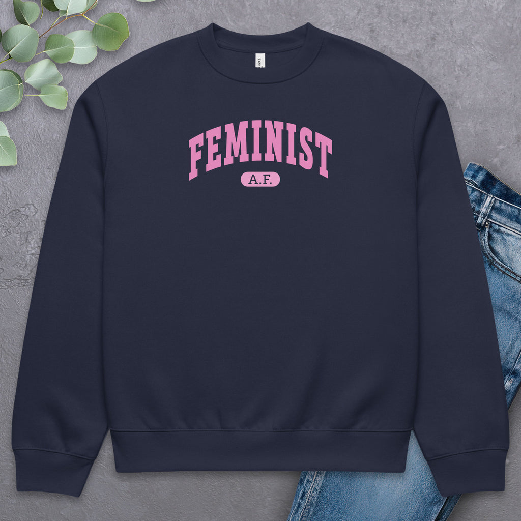 Flat navy Bella + Canvas heavyweight sweatshirt with pink text that reads “Feminist A.F.” in a collegiate style, styled with jeans and greenery.