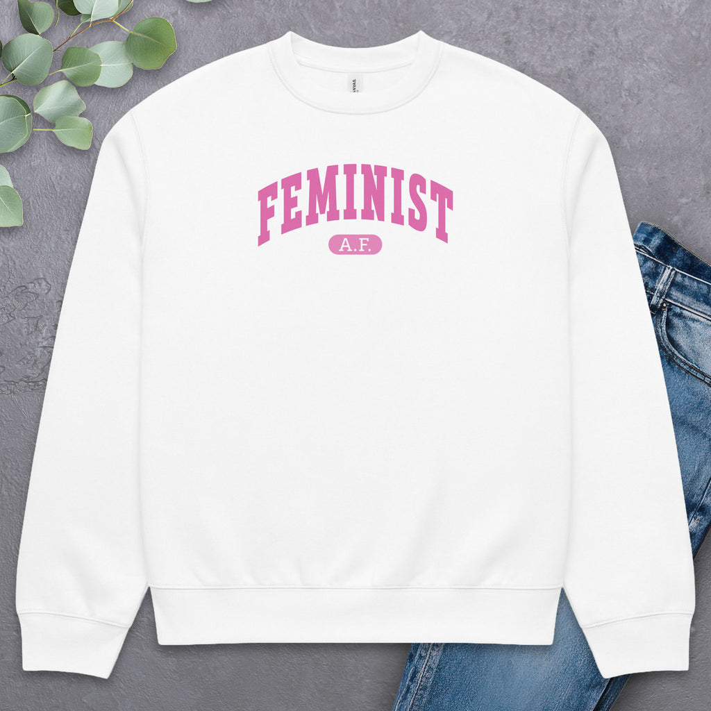 Flat white Bella + Canvas heavyweight sweatshirt with pink text that reads “Feminist A.F.” in a collegiate style, styled with jeans and greenery.