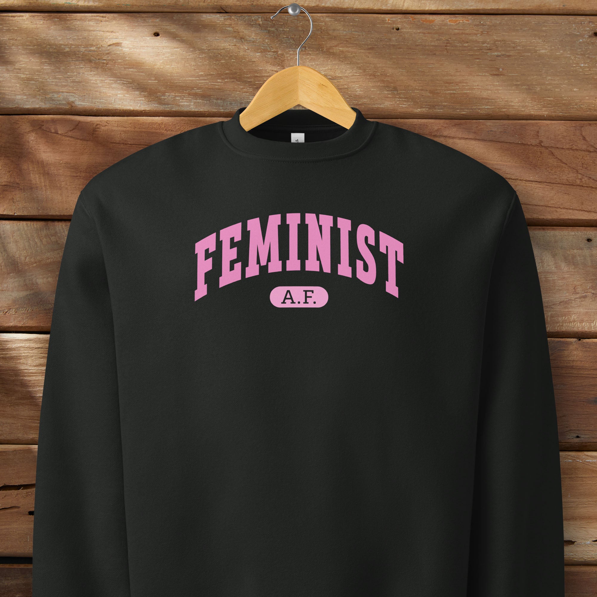 Hanging black Bella + Canvas heavyweight sweatshirt with pink text that reads “Feminist A.F.” in a collegiate style.