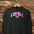 Hanging black Bella + Canvas heavyweight sweatshirt with pink text that reads “Feminist A.F.” in a collegiate style.