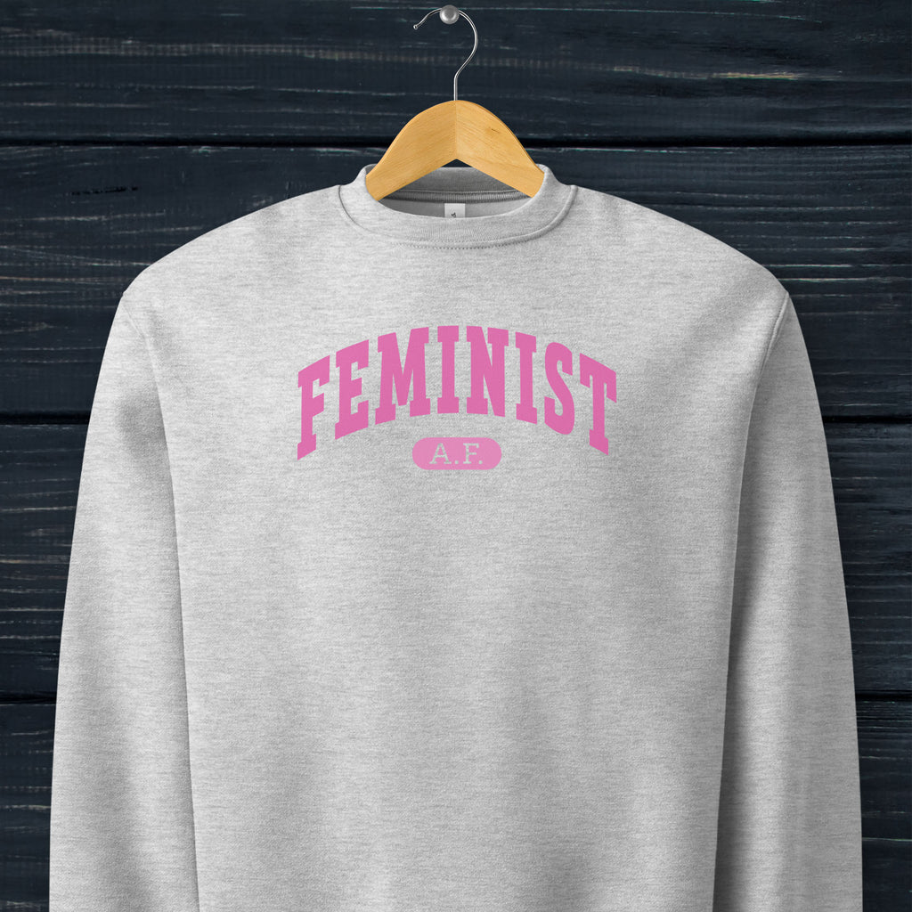 Hanging grey Bella + Canvas heavyweight sweatshirt with pink text that reads “Feminist A.F.” in a collegiate style.