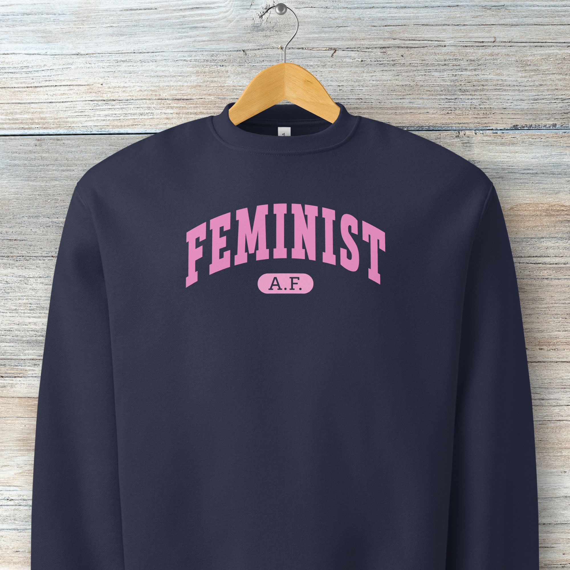 Hanging navy Bella + Canvas heavyweight sweatshirt with pink text that reads “Feminist A.F.” in a collegiate style.