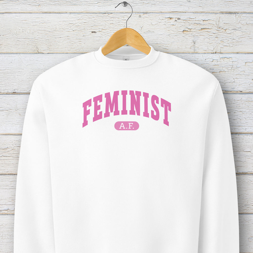 Hanging white Bella + Canvas heavyweight sweatshirt with pink text that reads “Feminist A.F.” in a collegiate style.