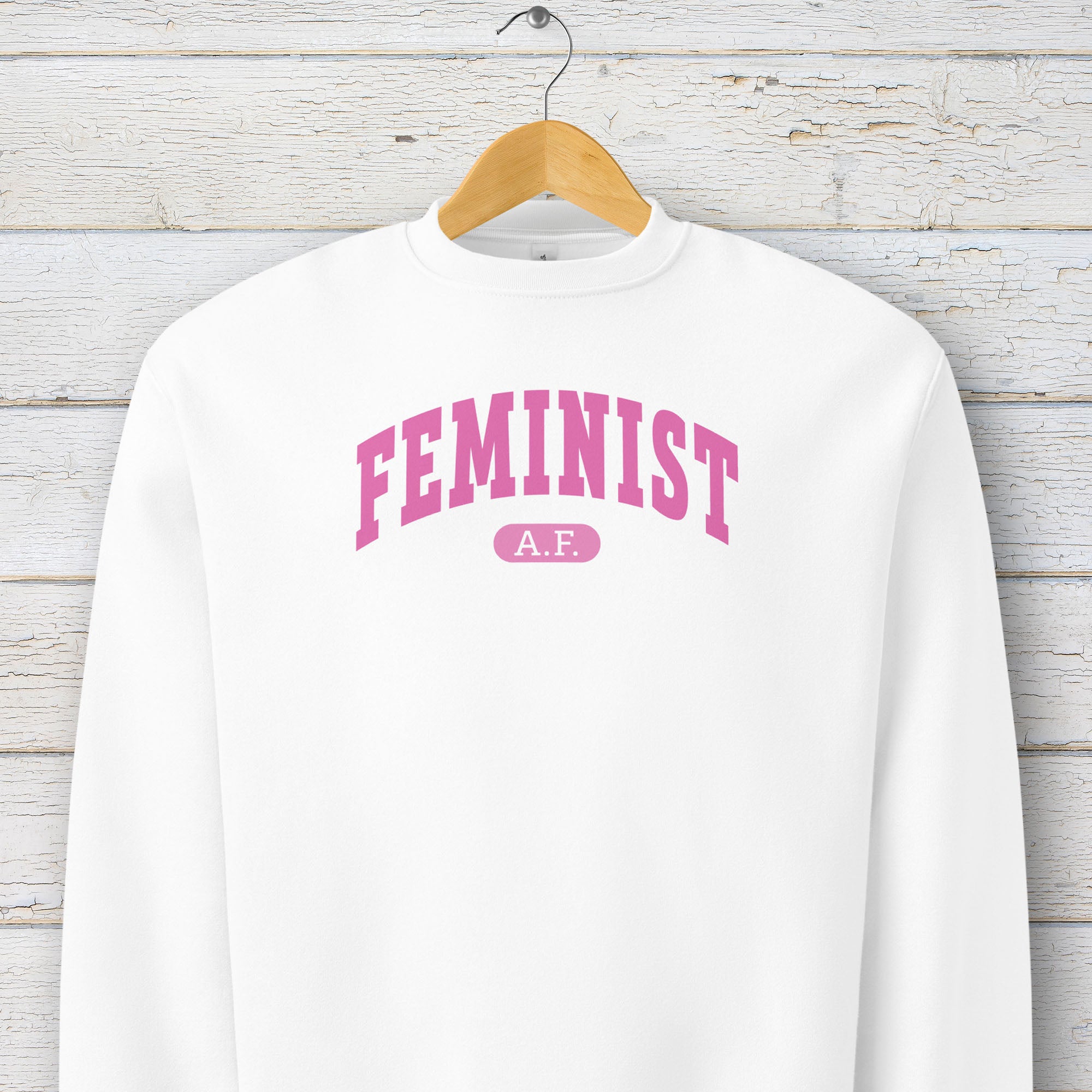 Hanging white Bella + Canvas heavyweight sweatshirt with pink text that reads “Feminist A.F.” in a collegiate style.