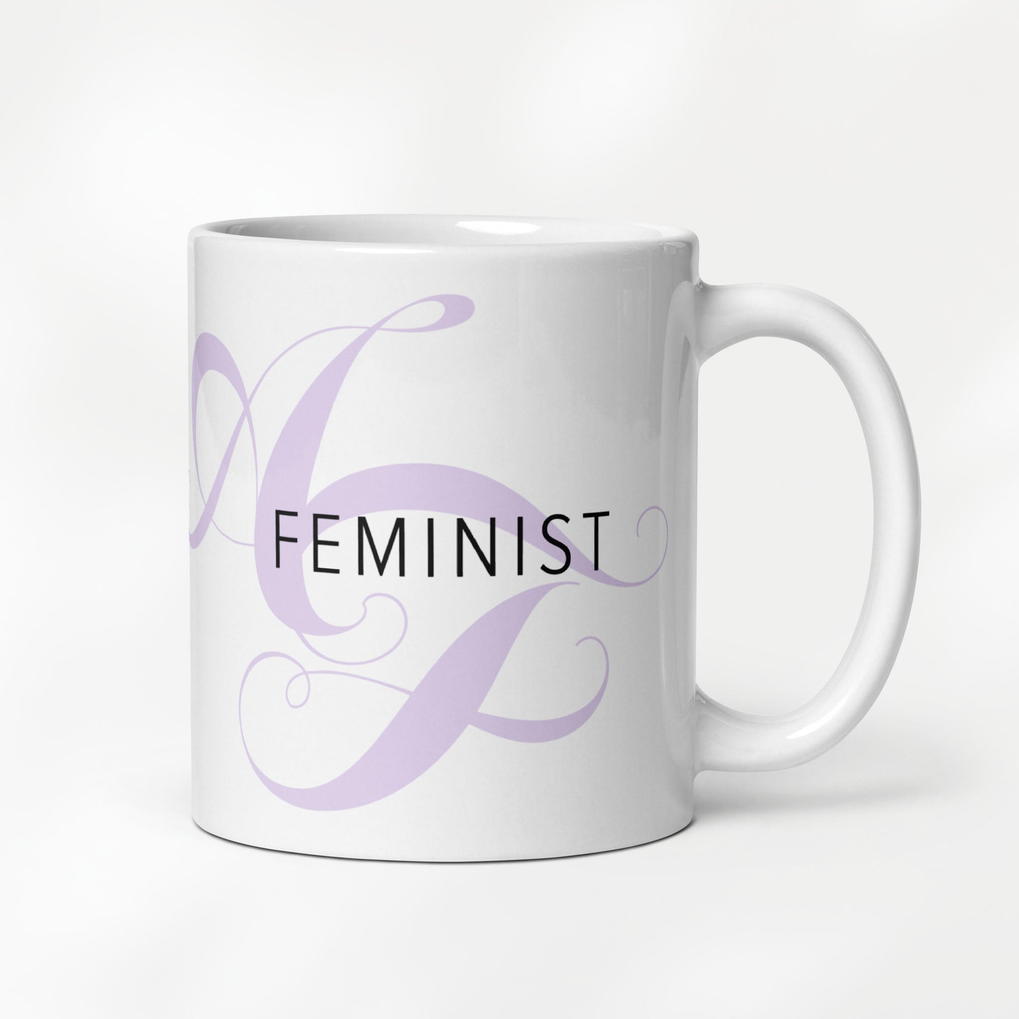 11 ounce white ceramic mug with the word “Feminist” in black text and a subtle lavender script design of the letters A and F in the background.