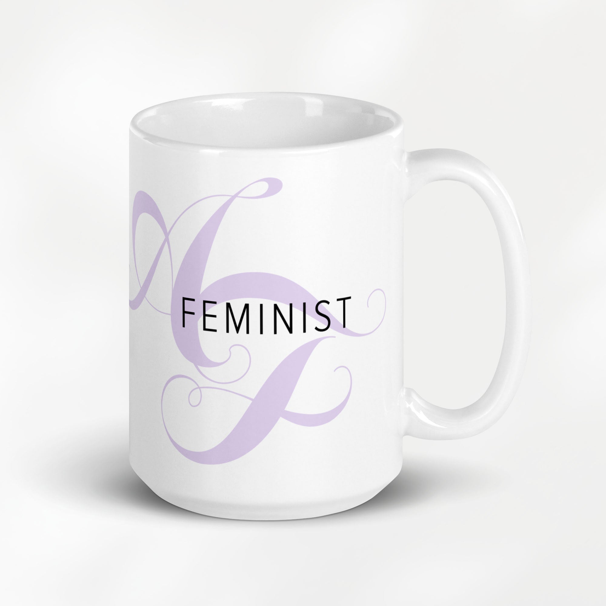 15 ounce white ceramic mug with the word “Feminist” in black text and a subtle lavender script design of the letters A and F in the background.