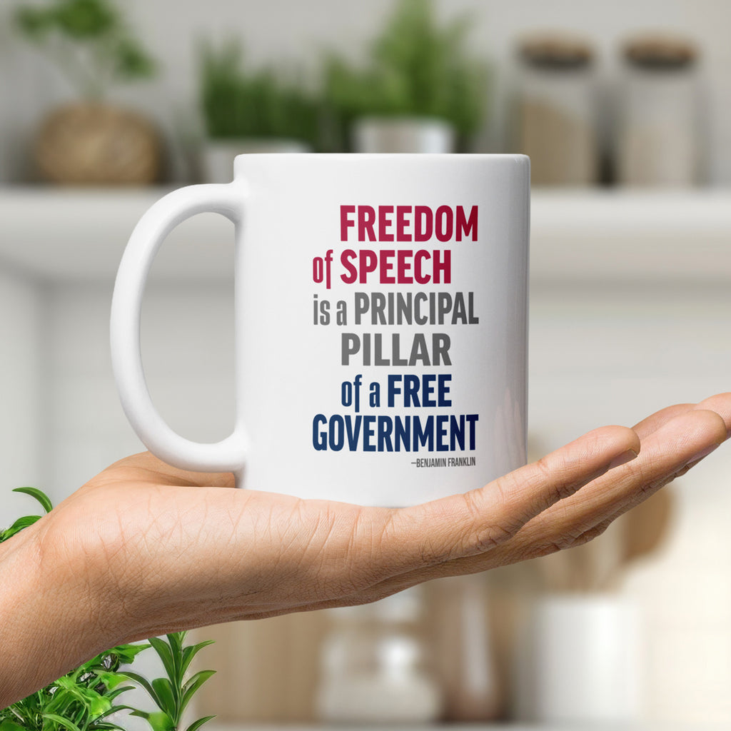 White ceramic mug with bold patriotic red, blue, and grey text that reads "Freedom of speech is a principal pillar of a free government. – Ben Franklin." Resting on a person's hand in a home.
