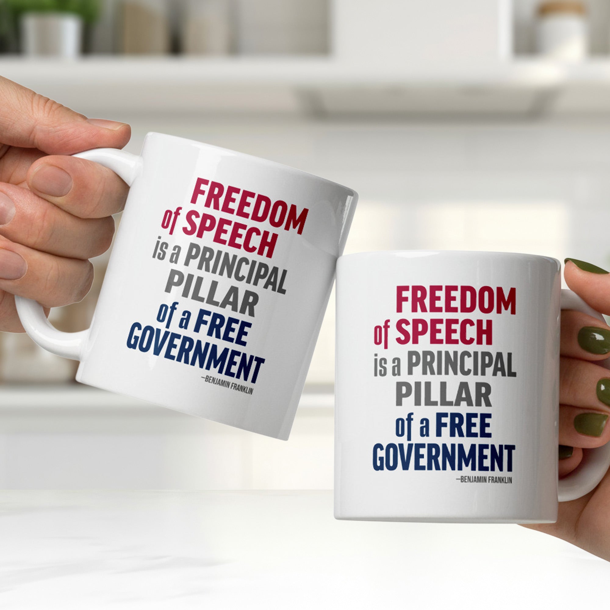Two people clinking white ceramic mugs in a kitchen, with mugs that have with bold patriotic red, blue, and grey text that reads "Freedom of speech is a principal pillar of a free government. – Ben Franklin."