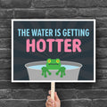 Downloadable, printable anti-trump protest sign with bold text in blue and pink that says "The water is getting hotter" above a cartoon illustration of a panicked frog in a pot of water.