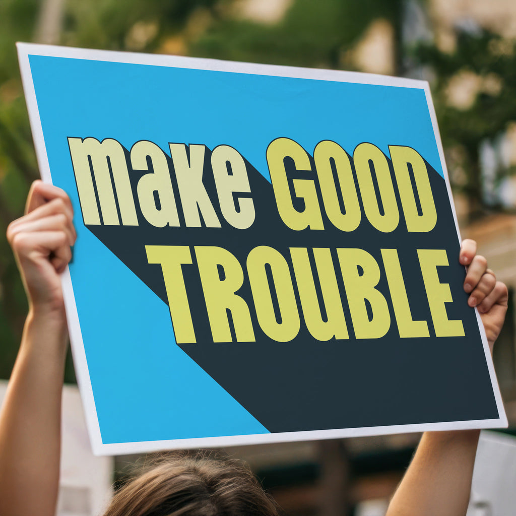 Person holding high above a crowd, a protest sign with bold text reading "Make Good Trouble" – a tribute to John Lewis’ legacy and a call for courageous, principled resistance against injustice.