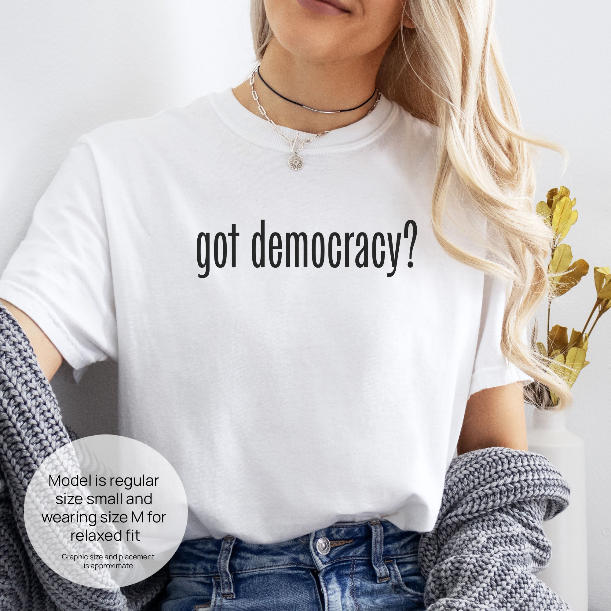 Female model wearing white t-shirt with white text reading “got democracy?” in minimalist font, styled like the classic “got milk?” design. Includes size and fit information for the model who is regular size small, and wearing a size M shirt for a relaxed fit.
