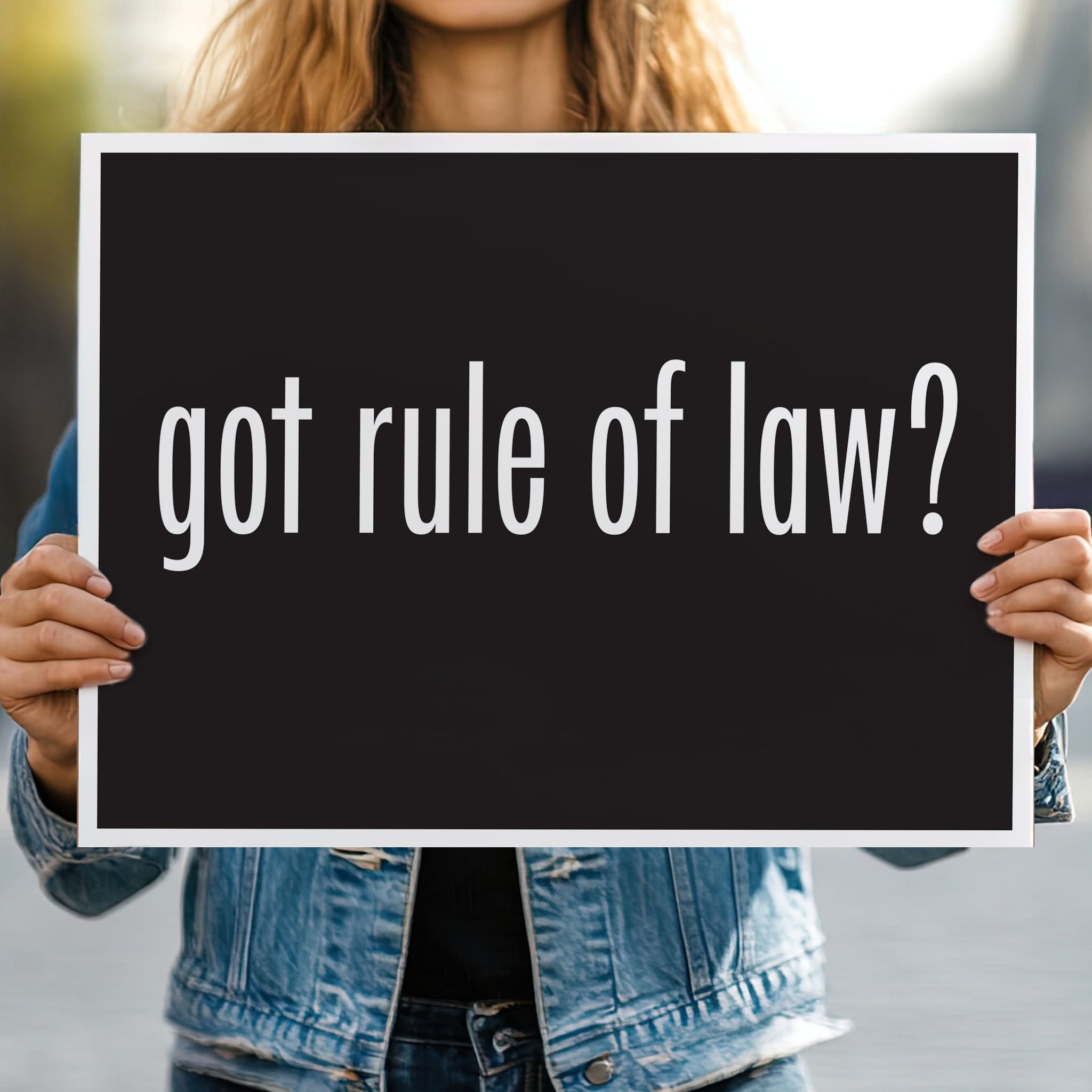 Person holding protest sign with black background and bold white text that says “got rule of law?” in the style of the “got milk?” campaign.