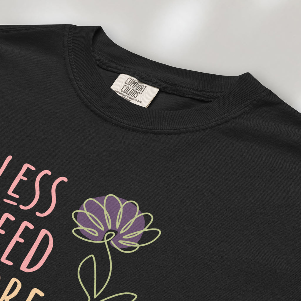 Closeup detail of black t-shirt featuring the phrase “Less Greed More Humanity” in friendly typography with a simple yellow flower illustration.