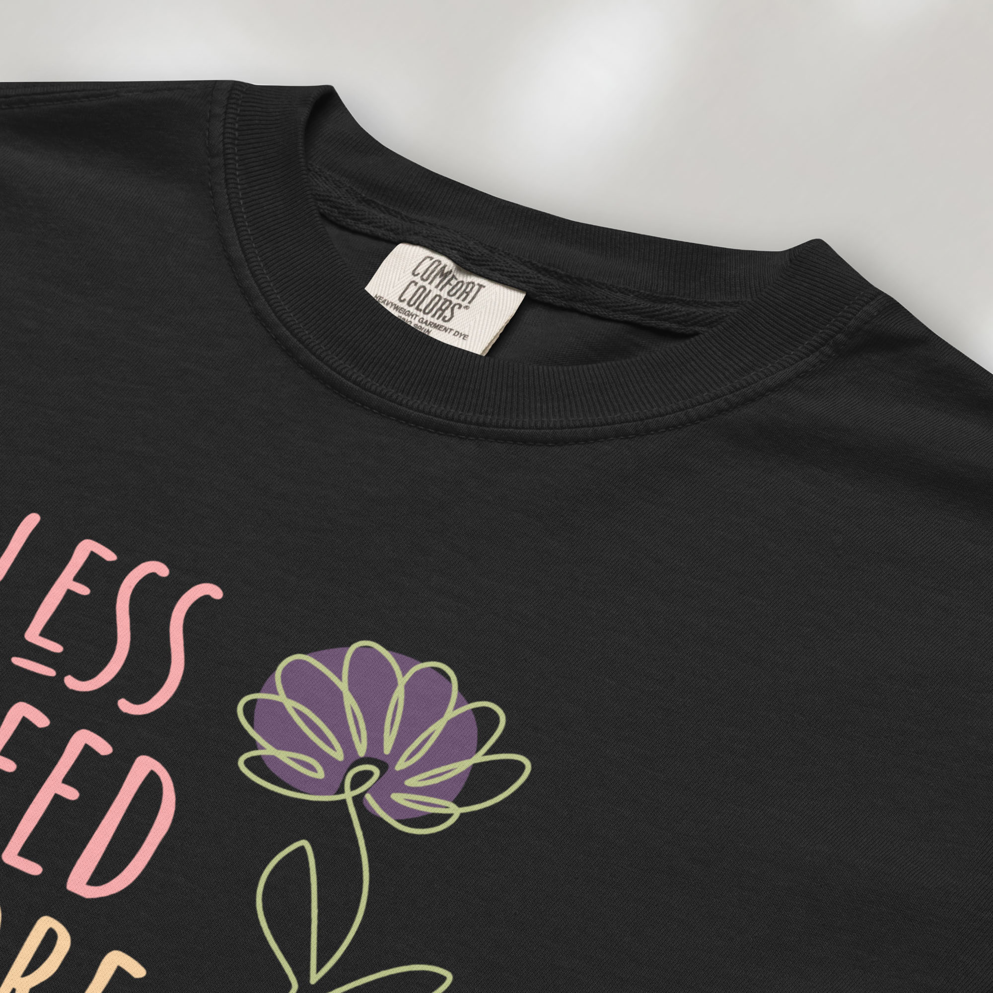 Closeup detail of black t-shirt featuring the phrase “Less Greed More Humanity” in friendly typography with a simple yellow flower illustration.