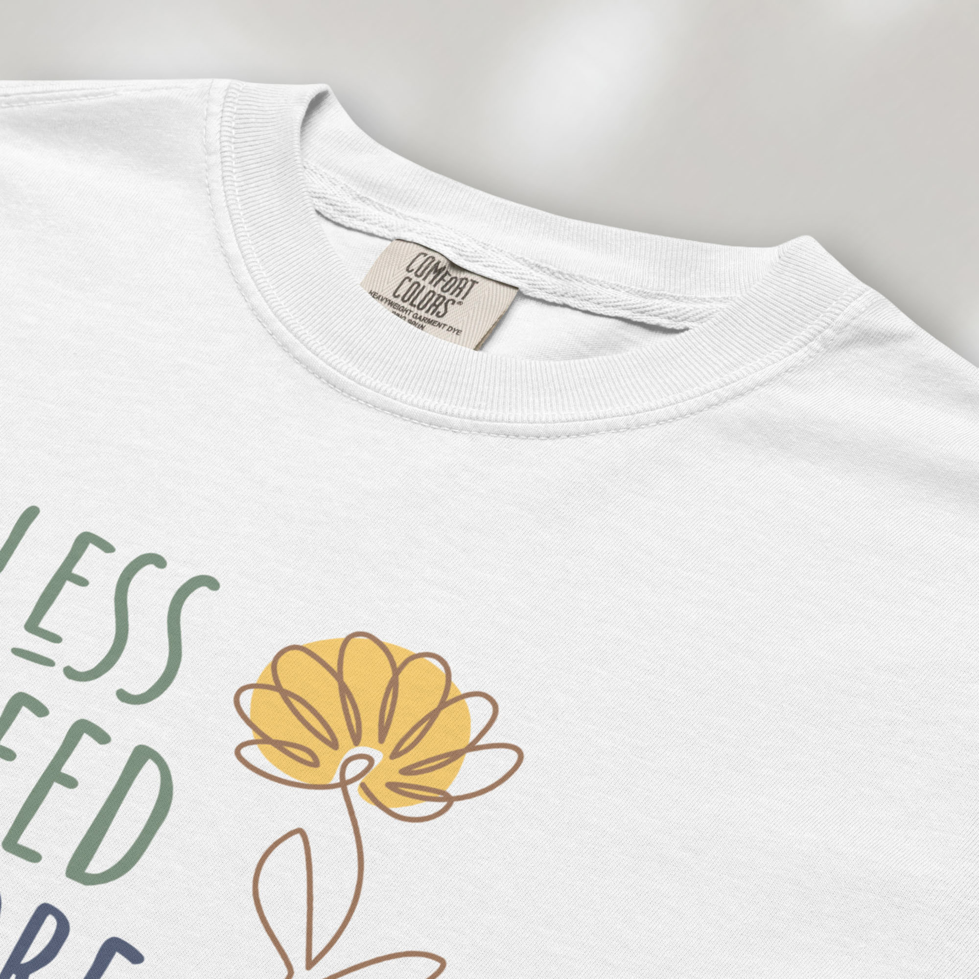 Closeup detail of white t-shirt featuring the phrase “Less Greed More Humanity” in friendly typography with a simple yellow flower illustration.