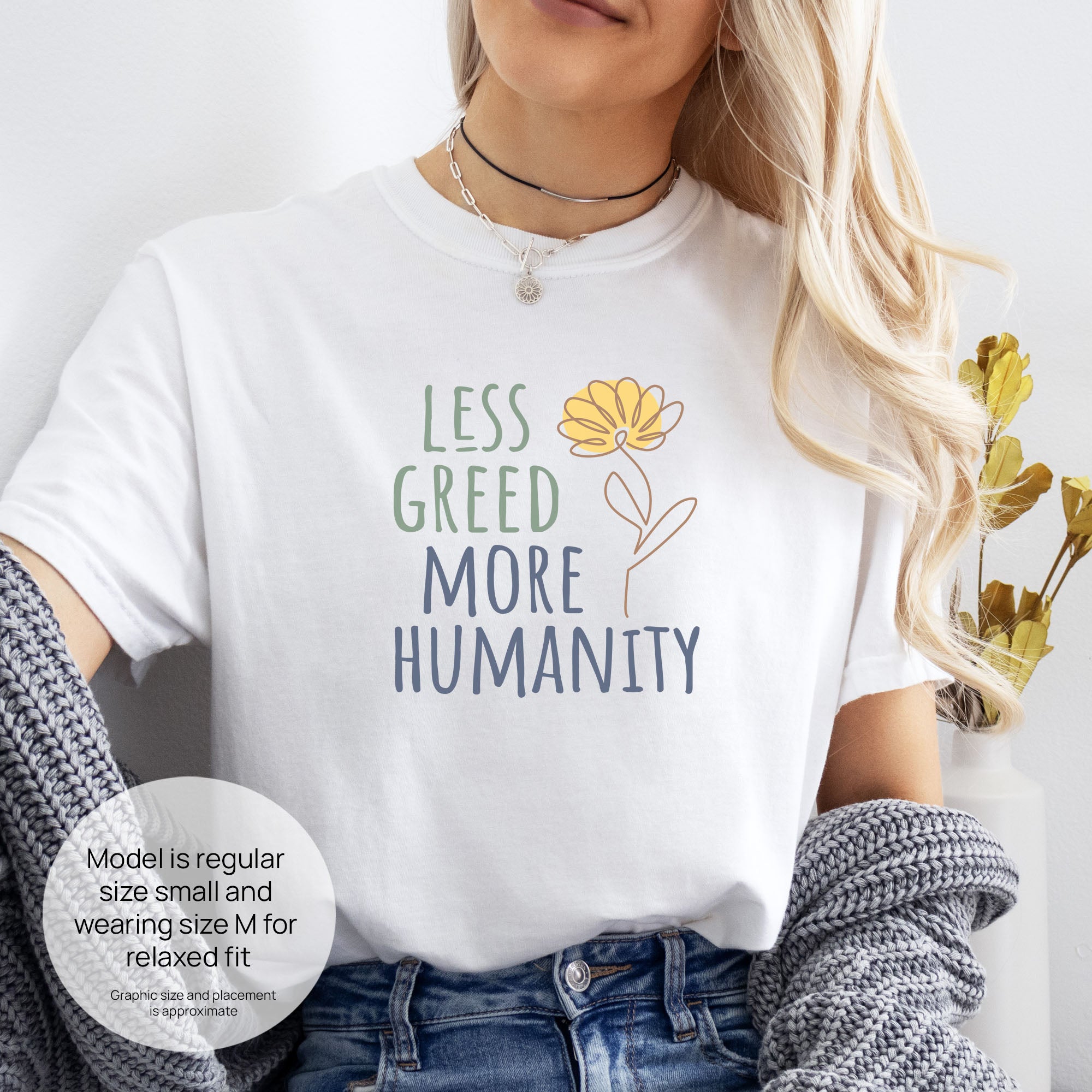 Female model wearing white t-shirt featuring the phrase “Less Greed More Humanity” in friendly typography with a simple yellow flower illustration. Includes size and fit information for the model who is regular size small, and wearing a size M shirt for a relaxed fit.
