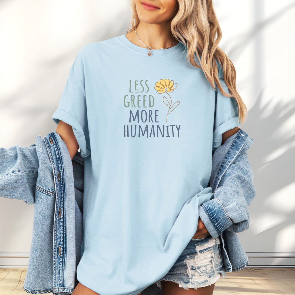Female model wearing oversized chambray t-shirt featuring the phrase “Less Greed More Humanity” in friendly typography with a simple yellow flower illustration.