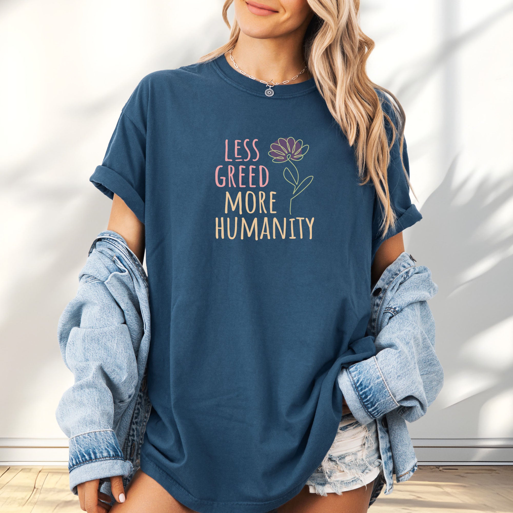 Female model wearing oversized midnight t-shirt featuring the phrase “Less Greed More Humanity” in friendly typography with a simple yellow flower illustration.