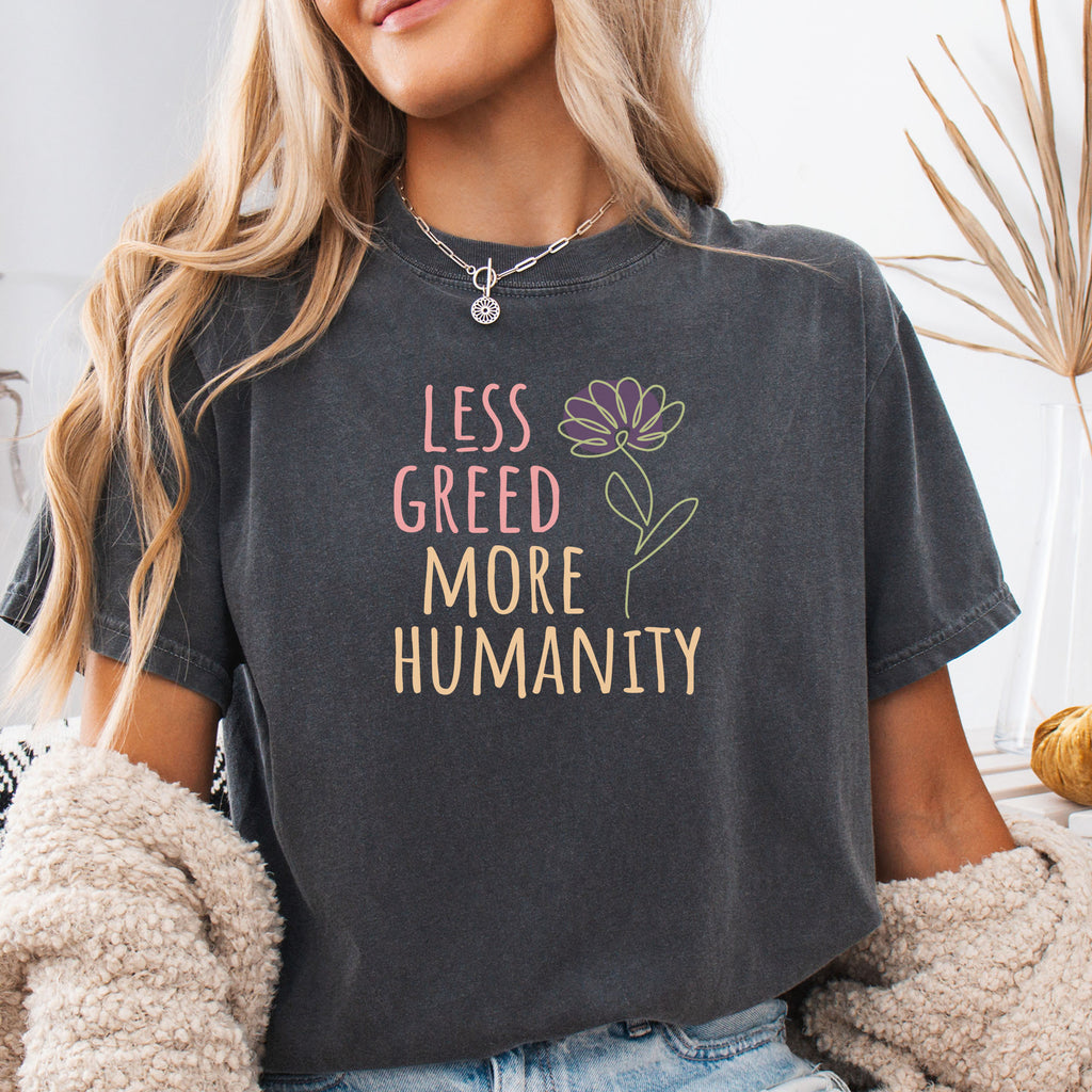Female model wearing pepper t-shirt featuring the phrase “Less Greed More Humanity” in friendly typography with a simple yellow flower illustration.