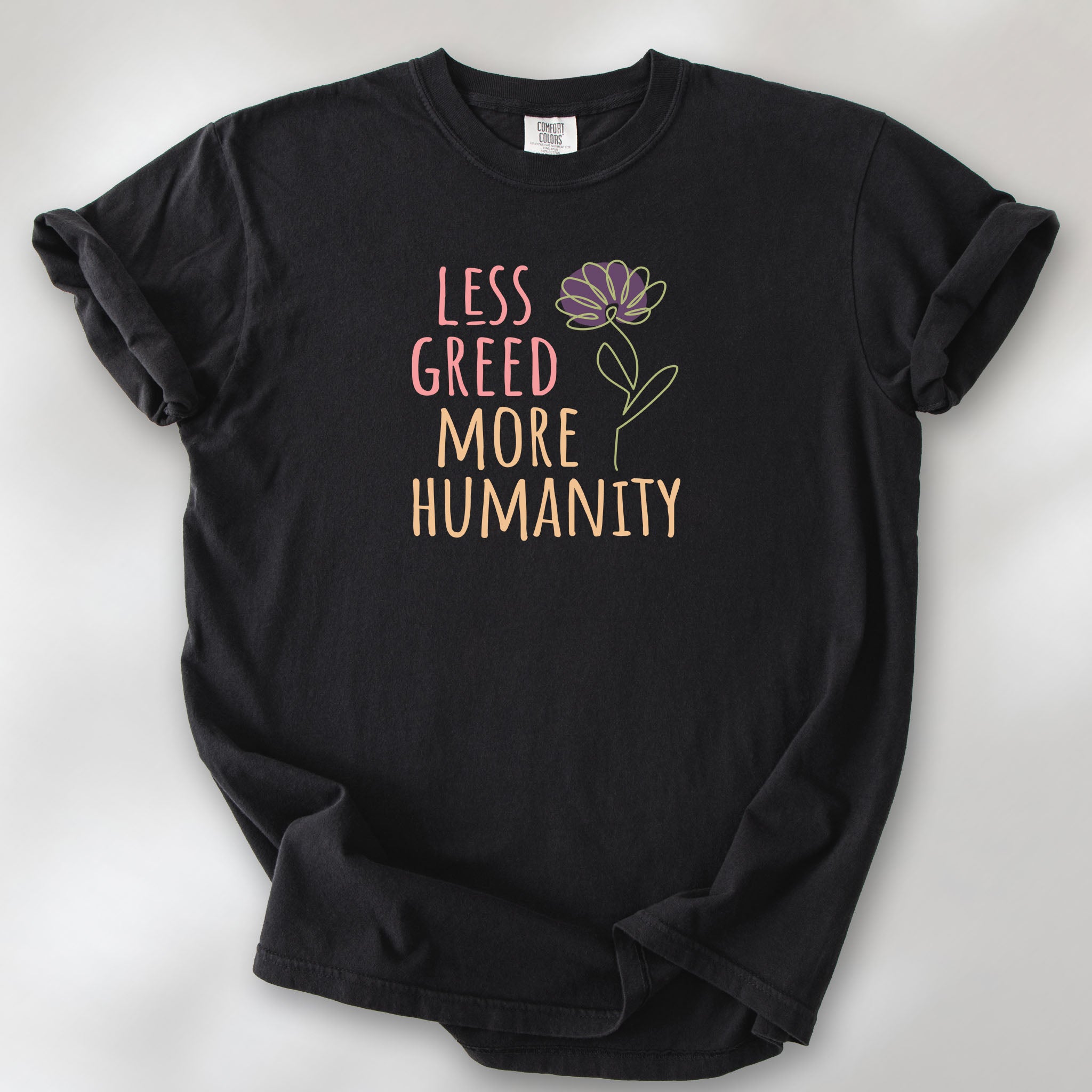 Black t-shirt featuring the phrase “Less Greed More Humanity” in friendly typography with a simple yellow flower illustration.