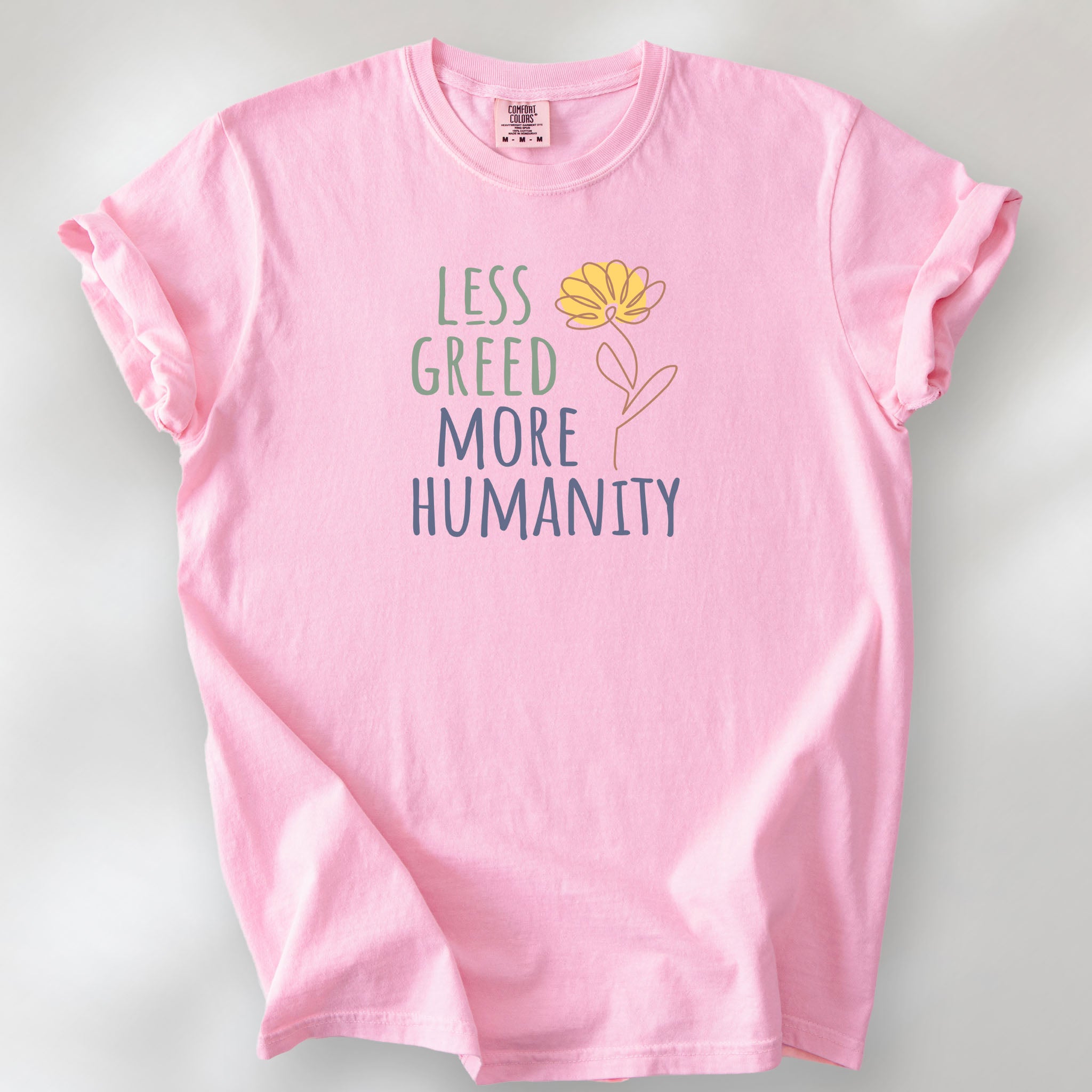 Blossom t-shirt featuring the phrase “Less Greed More Humanity” in friendly typography with a simple yellow flower illustration.
