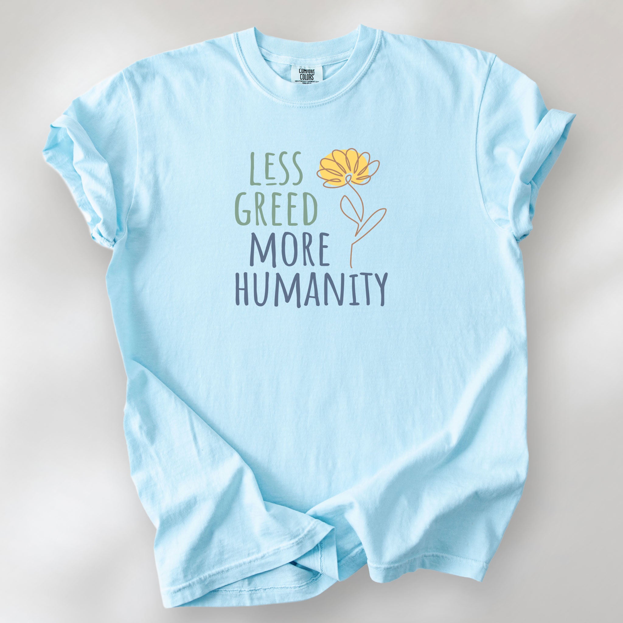Chambray t-shirt featuring the phrase “Less Greed More Humanity” in friendly typography with a simple yellow flower illustration.