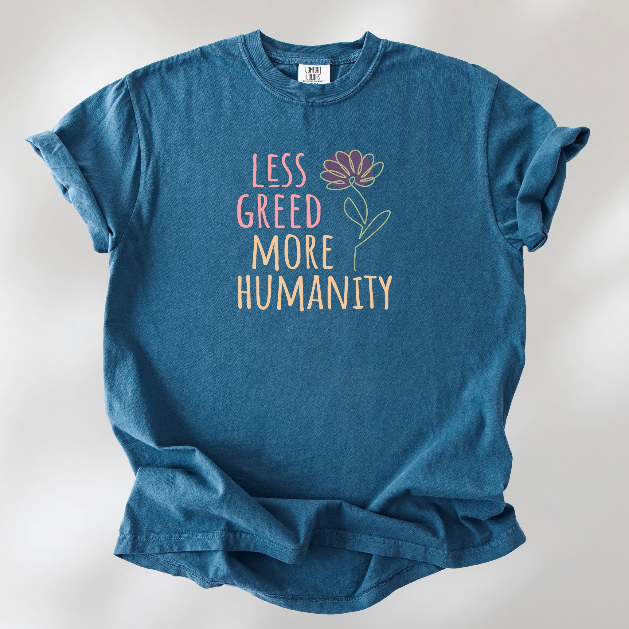 Midnight t-shirt featuring the phrase “Less Greed More Humanity” in friendly typography with a simple yellow flower illustration.