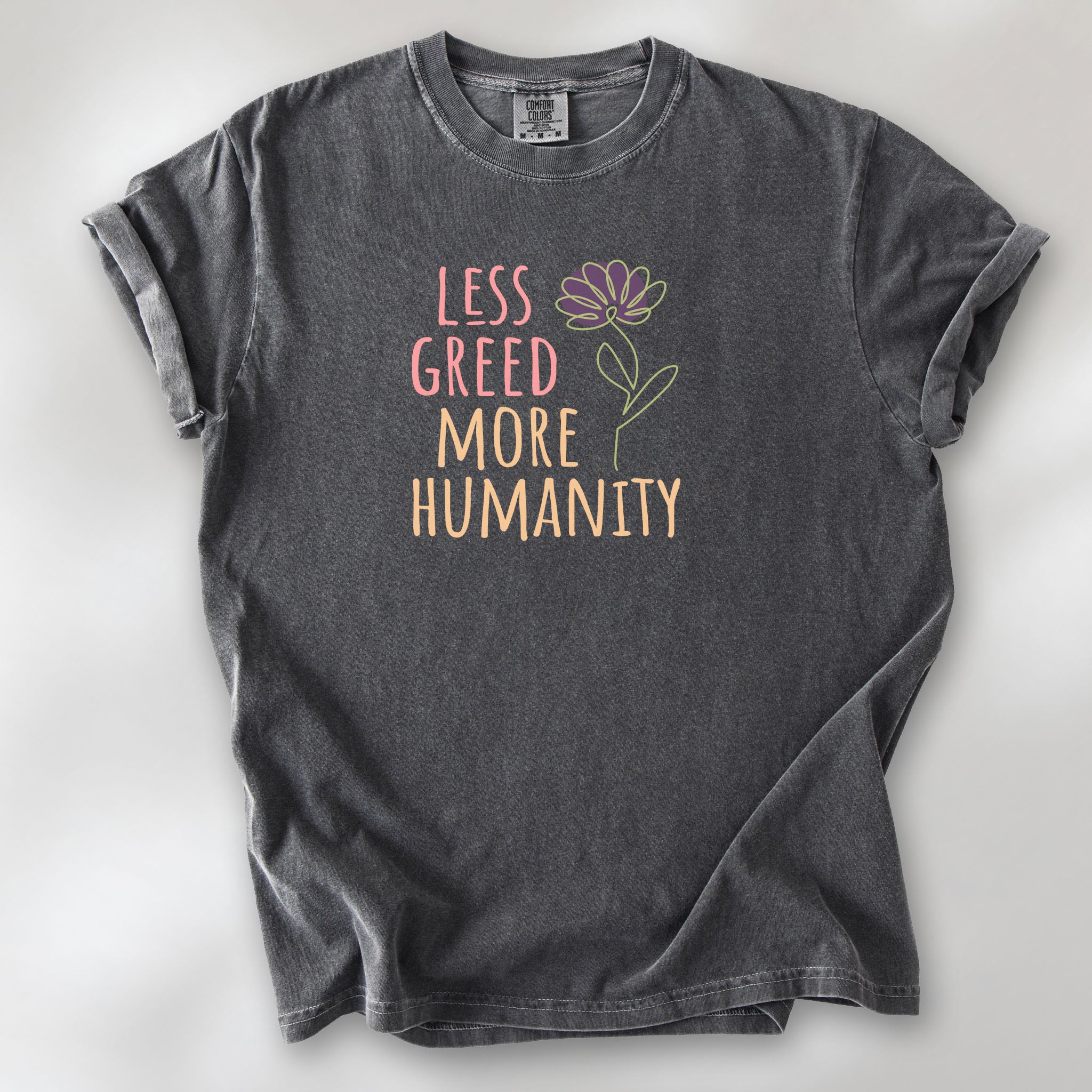 Pepper t-shirt featuring the phrase “Less Greed More Humanity” in friendly typography with a simple yellow flower illustration.