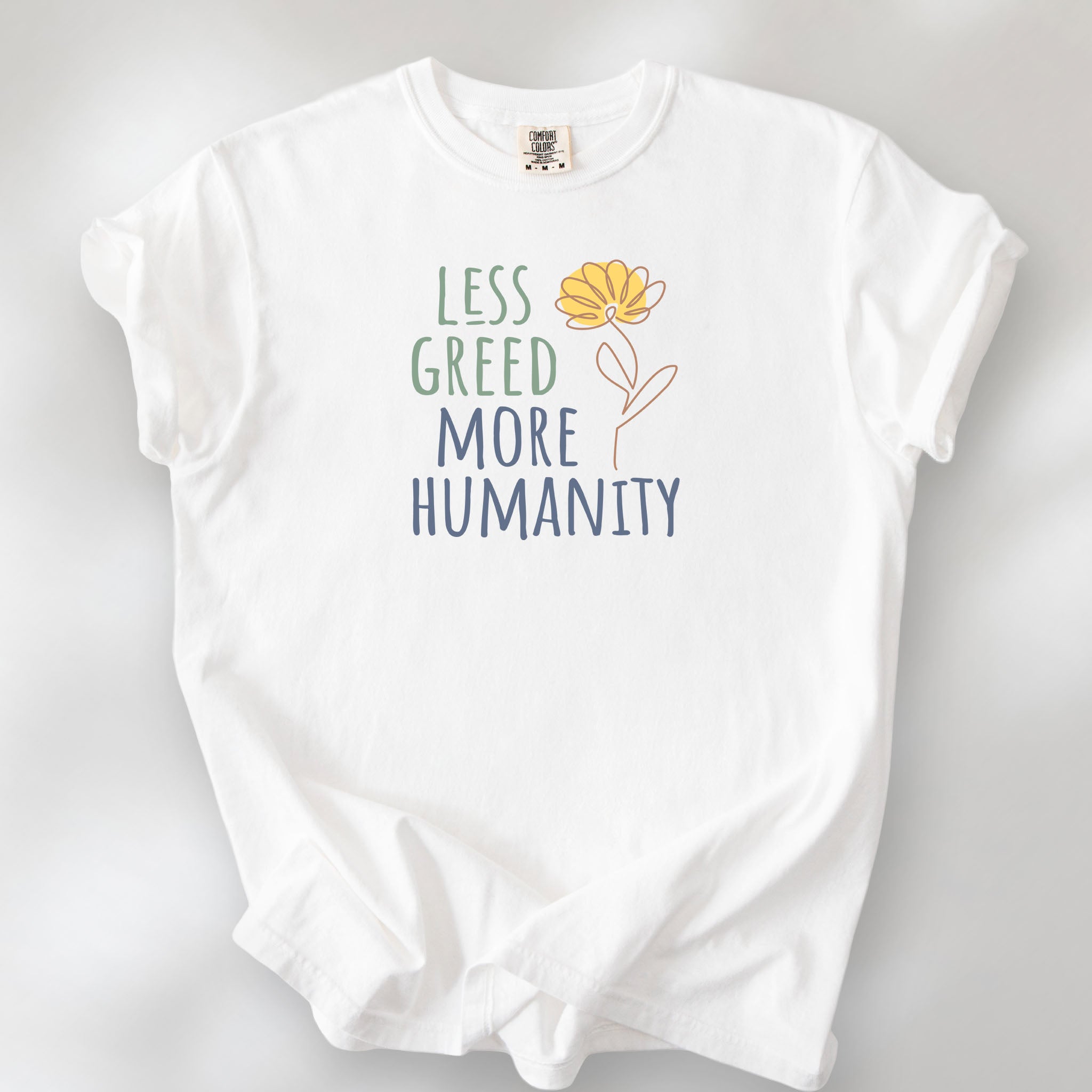 White t-shirt featuring the phrase “Less Greed More Humanity” in friendly typography with a simple yellow flower illustration.