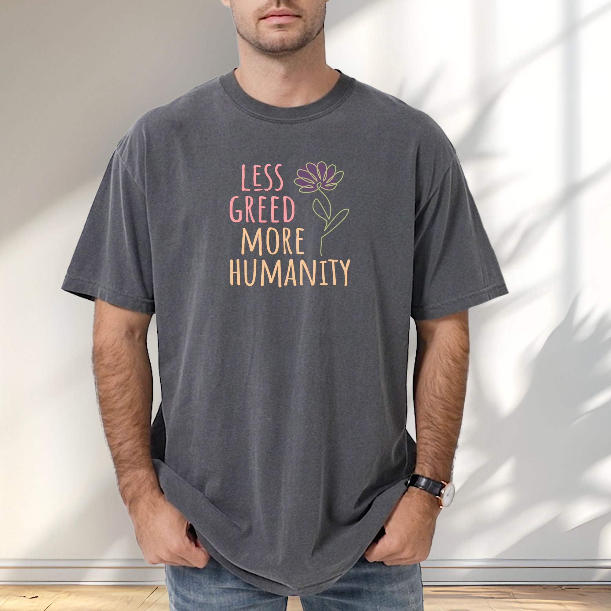 Male model wearing pepper t-shirt featuring the phrase “Less Greed More Humanity” in friendly typography with a simple yellow flower illustration.