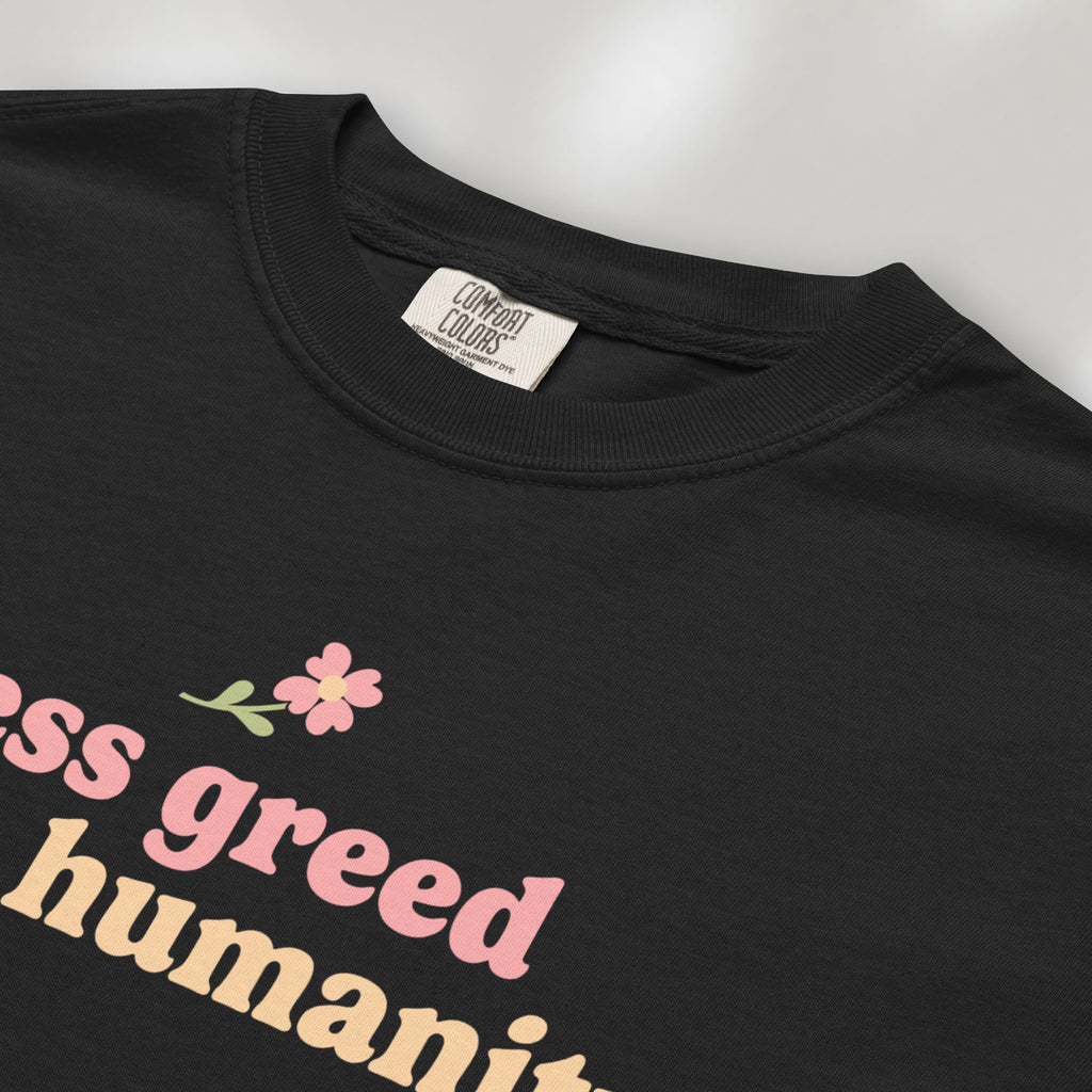 Closeup detail of black t-shirt featuring the phrase "Less Greed More Humanity" in soft retro typography with a small flower graphic above the text.