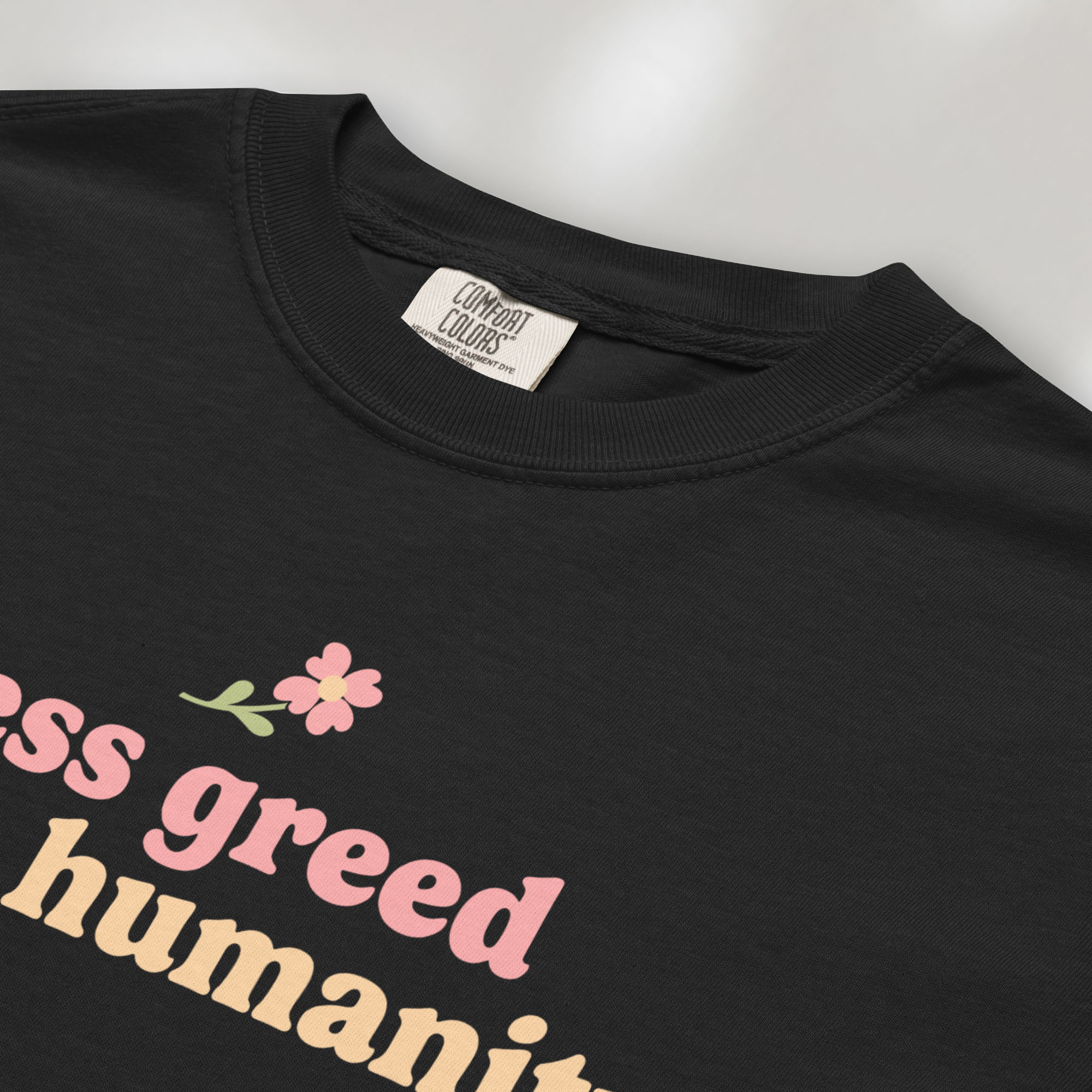 Closeup detail of black t-shirt featuring the phrase "Less Greed More Humanity" in soft retro typography with a small flower graphic above the text.