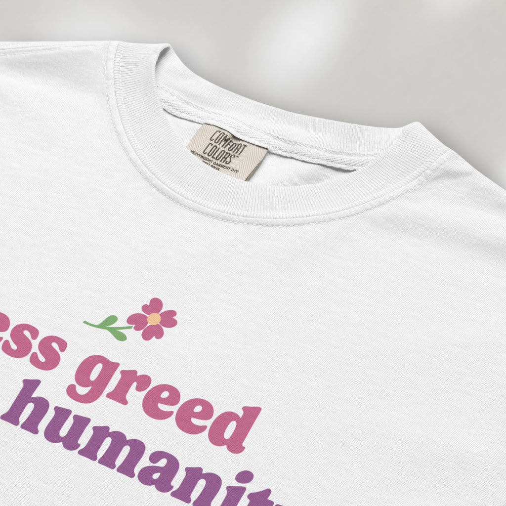 Closeup detail of white t-shirt featuring the phrase "Less Greed More Humanity" in soft retro typography with a small flower graphic above the text.