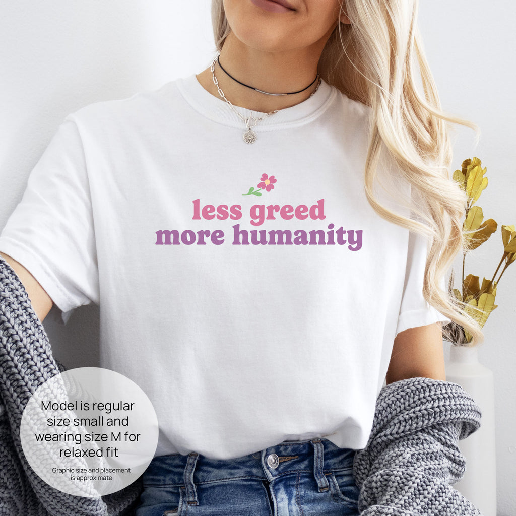 Female model wearing white t-shirt featuring the phrase "Less Greed More Humanity" in soft retro typography with a small flower graphic above the text. Includes size and fit information for the model who is regular size small, and wearing a size M shirt for a relaxed fit.
