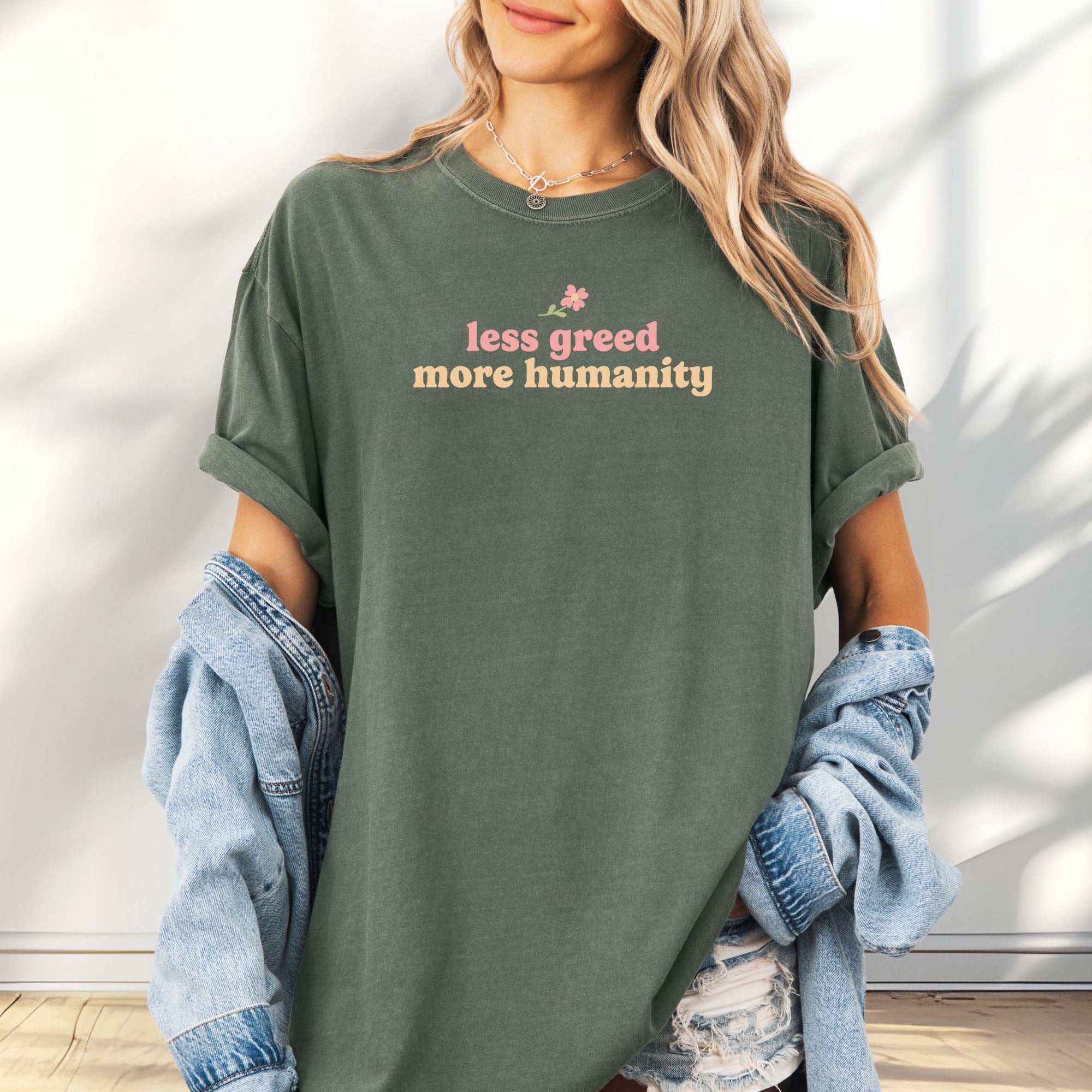 Female model wearing oversized moss t-shirt featuring the phrase "Less Greed More Humanity" in soft retro typography with a small flower graphic above the text.