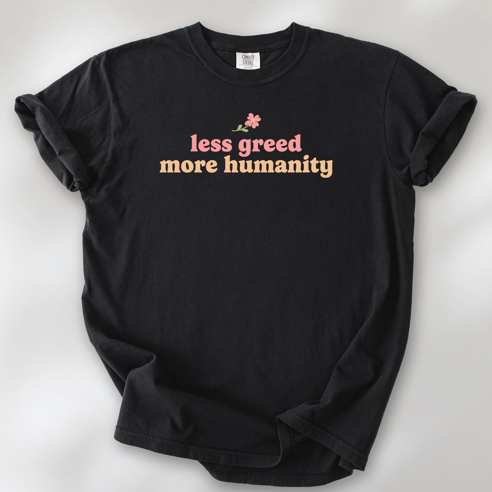 Black t-shirt featuring the phrase "Less Greed More Humanity" in soft retro typography with a small flower graphic above the text.