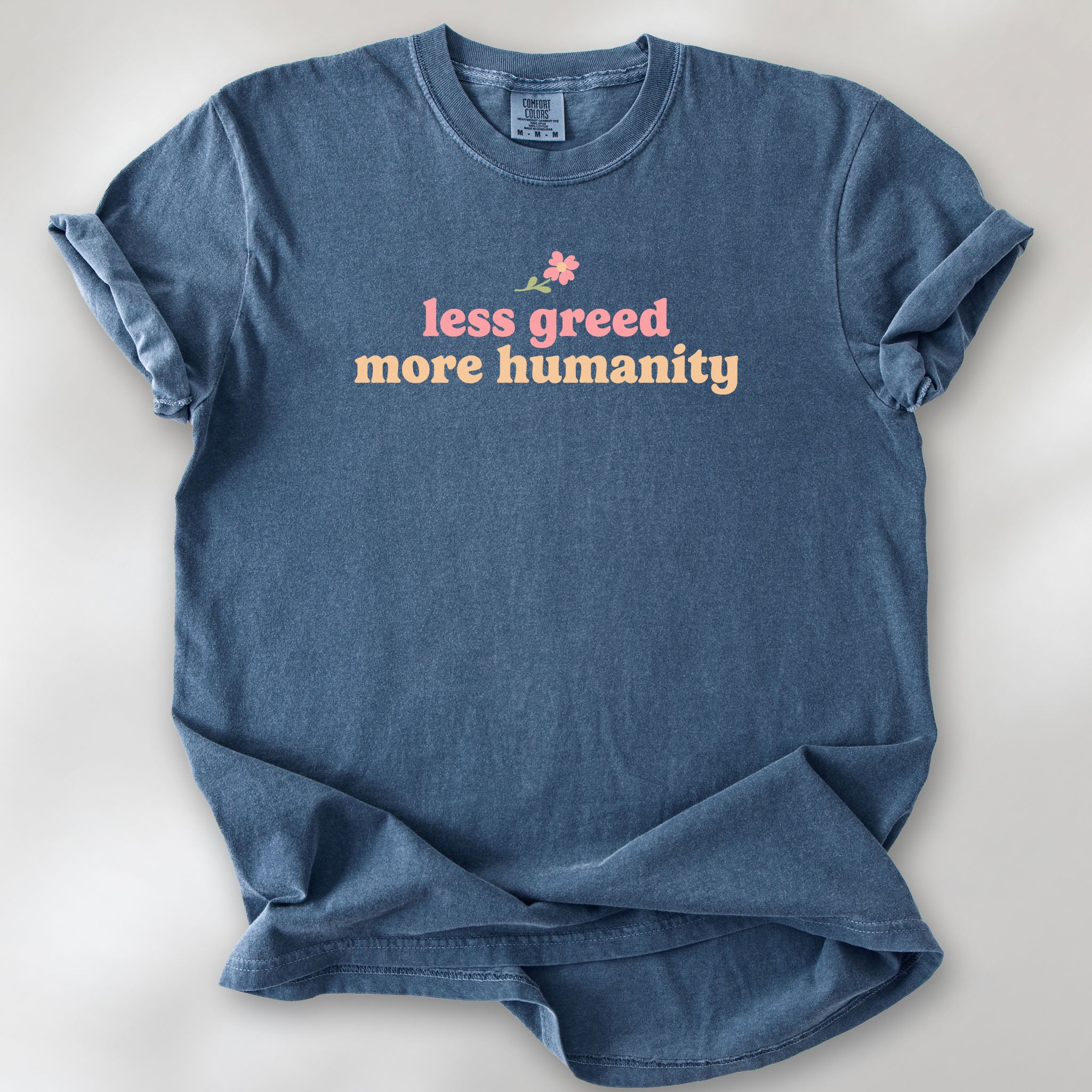 Denim t-shirt featuring the phrase "Less Greed More Humanity" in soft retro typography with a small flower graphic above the text.