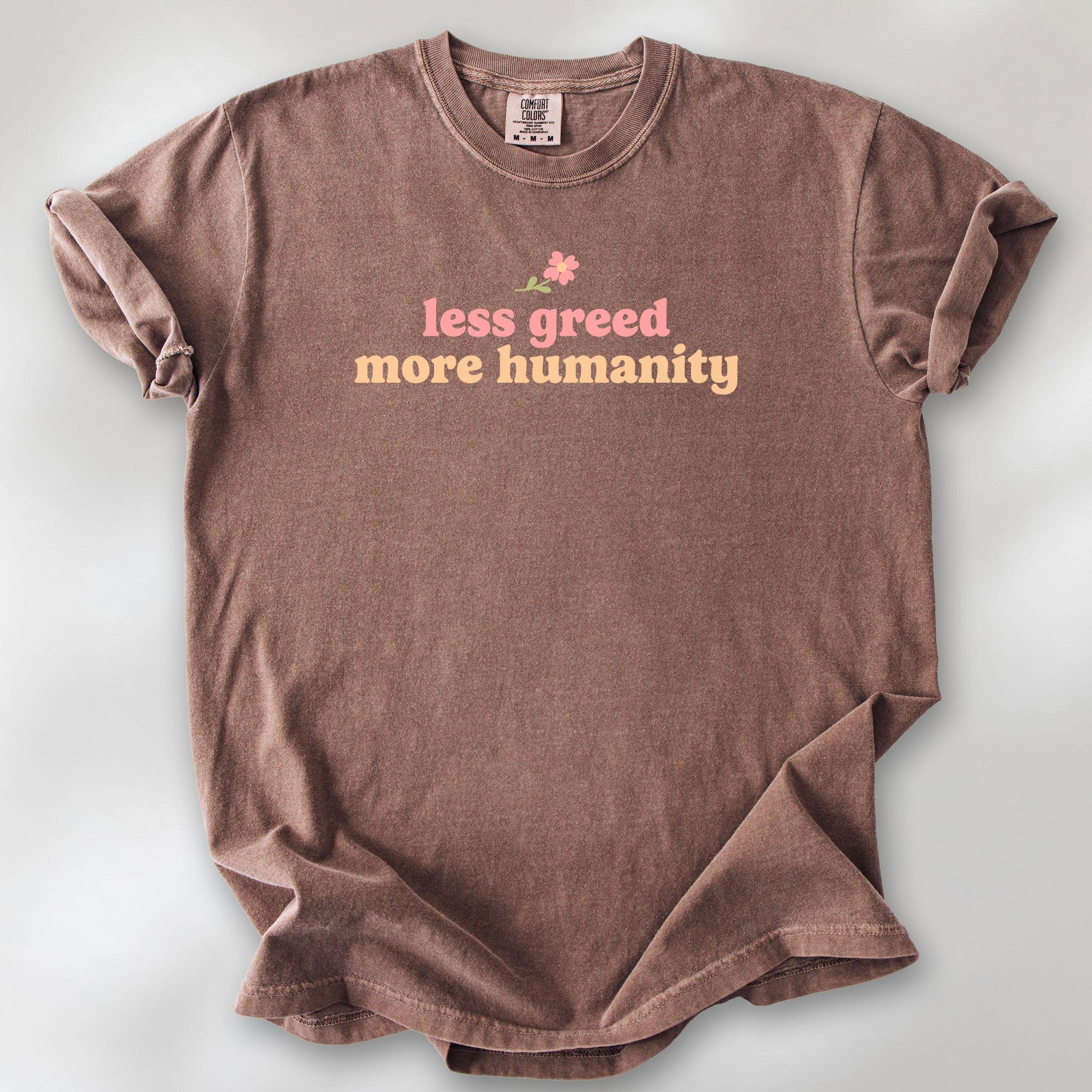 Espresso t-shirt featuring the phrase "Less Greed More Humanity" in soft retro typography with a small flower graphic above the text.