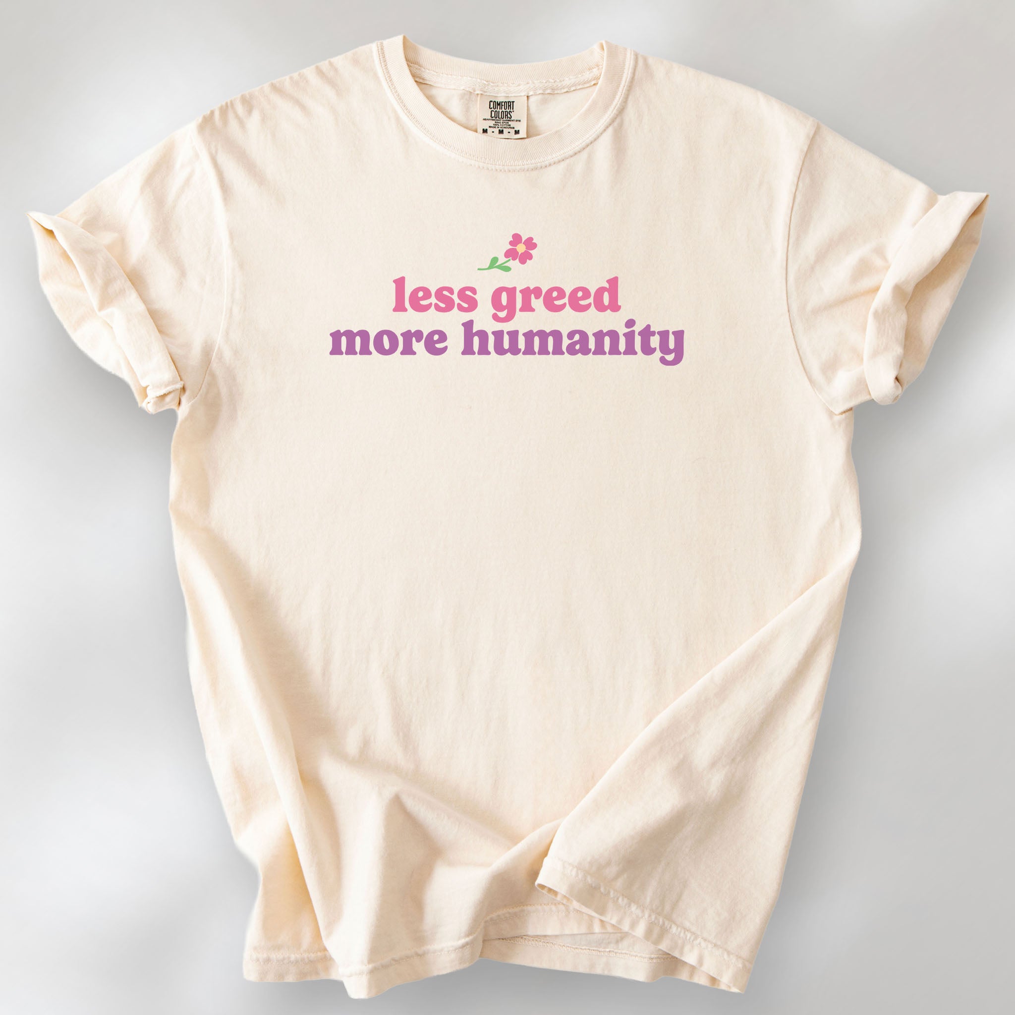 Ivory t-shirt featuring the phrase "Less Greed More Humanity" in soft retro typography with a small flower graphic above the text.